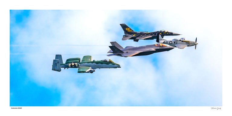 An A-10, F-35, F-16, and silver P-51 Mustang fly together toward the right against a blue sky filled with soft white clouds.