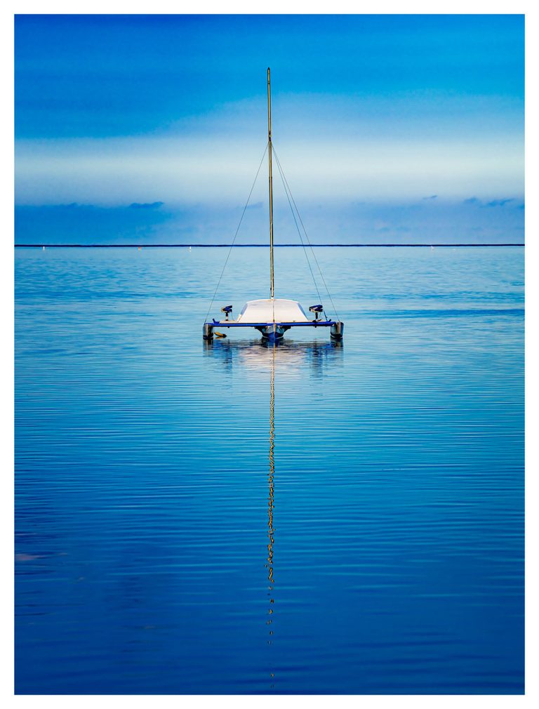 A small white sailboat with a bare mast floats on calm blue water, its long golden reflection extending toward the foreground beneath a band of pale clouds.