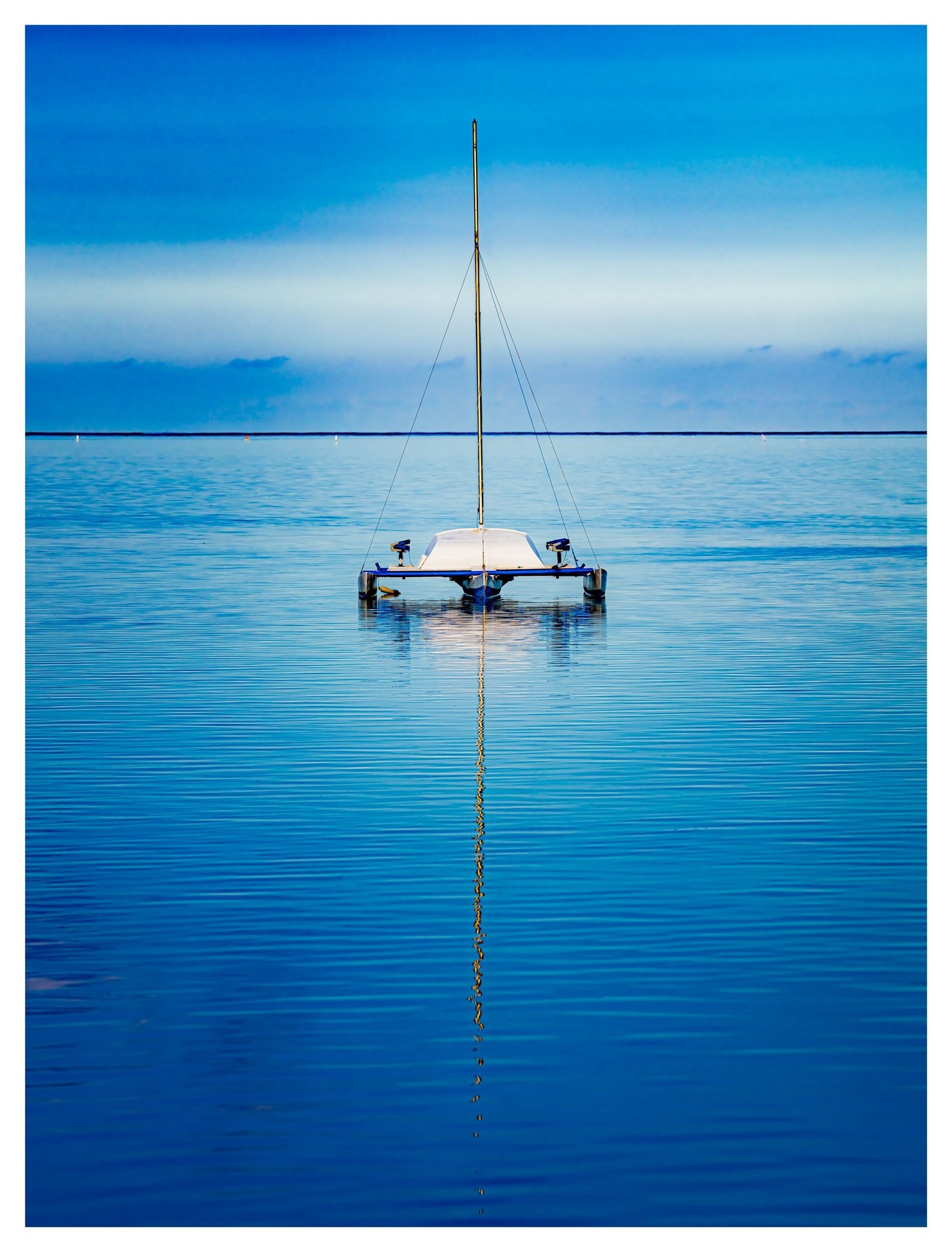 A small white sailboat with a bare mast floats on calm blue water, its long golden reflection extending toward the foreground beneath a band of pale clouds.