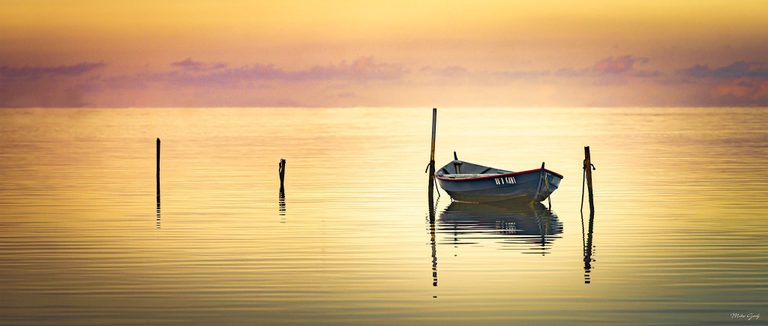 A small blue boat with red trim floats among mooring posts on calm golden water beneath a softly clouded orange sky.