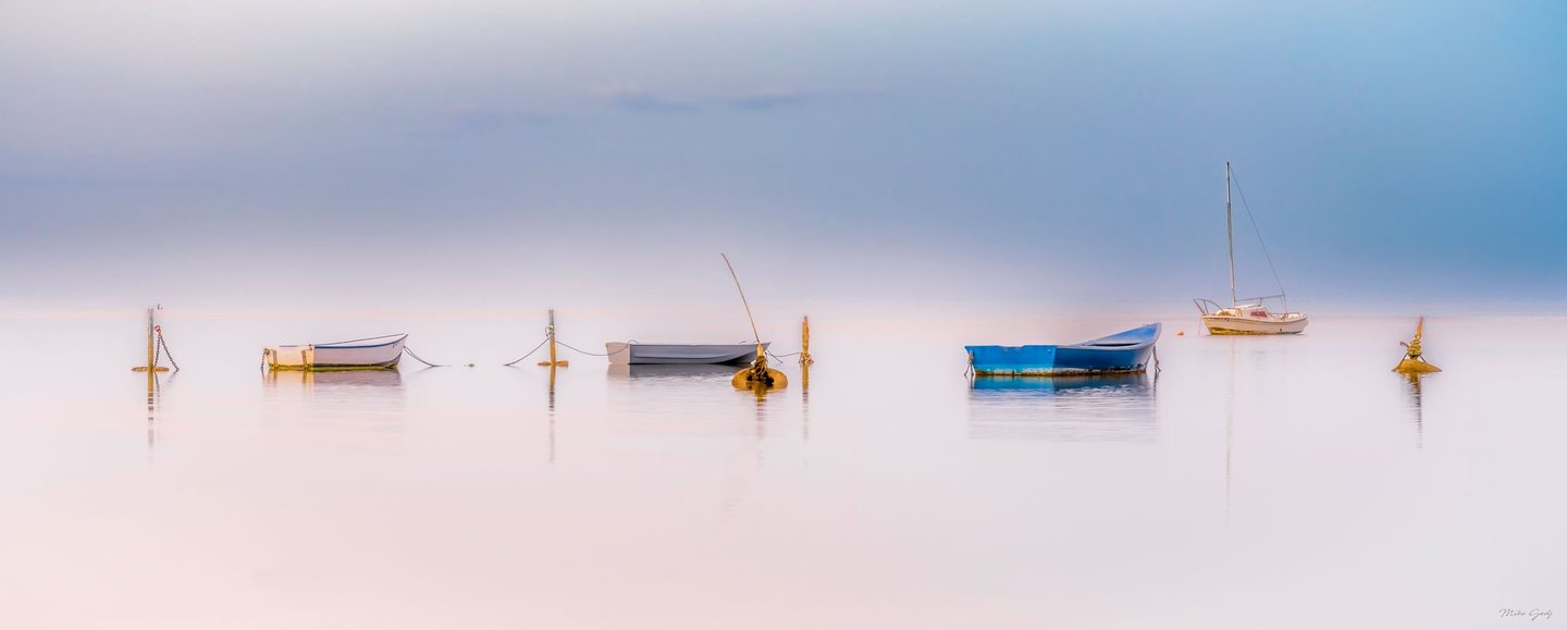 Three small rowboats and a sailboat rest among mooring posts on calm pink-tinted water beneath a hazy blue sky.