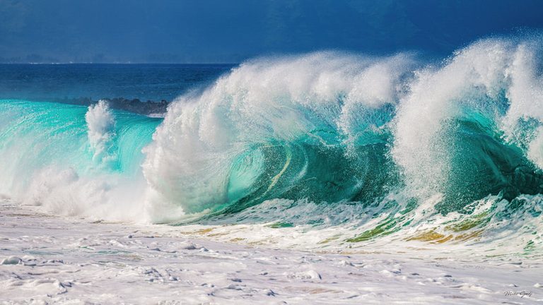Large turquoise ocean waves break into white spray above a foam-covered foreground, with dark blue water and a hazy mountainous coastline behind them.