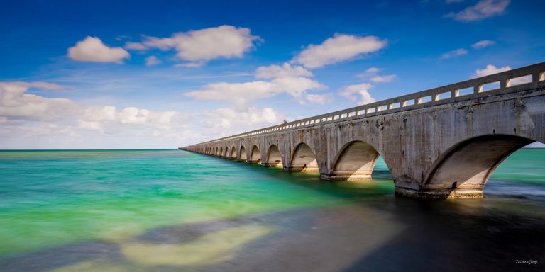 A long concrete bridge with repeating arches stretches toward the horizon over turquoise water beneath a blue sky scattered with white clouds.