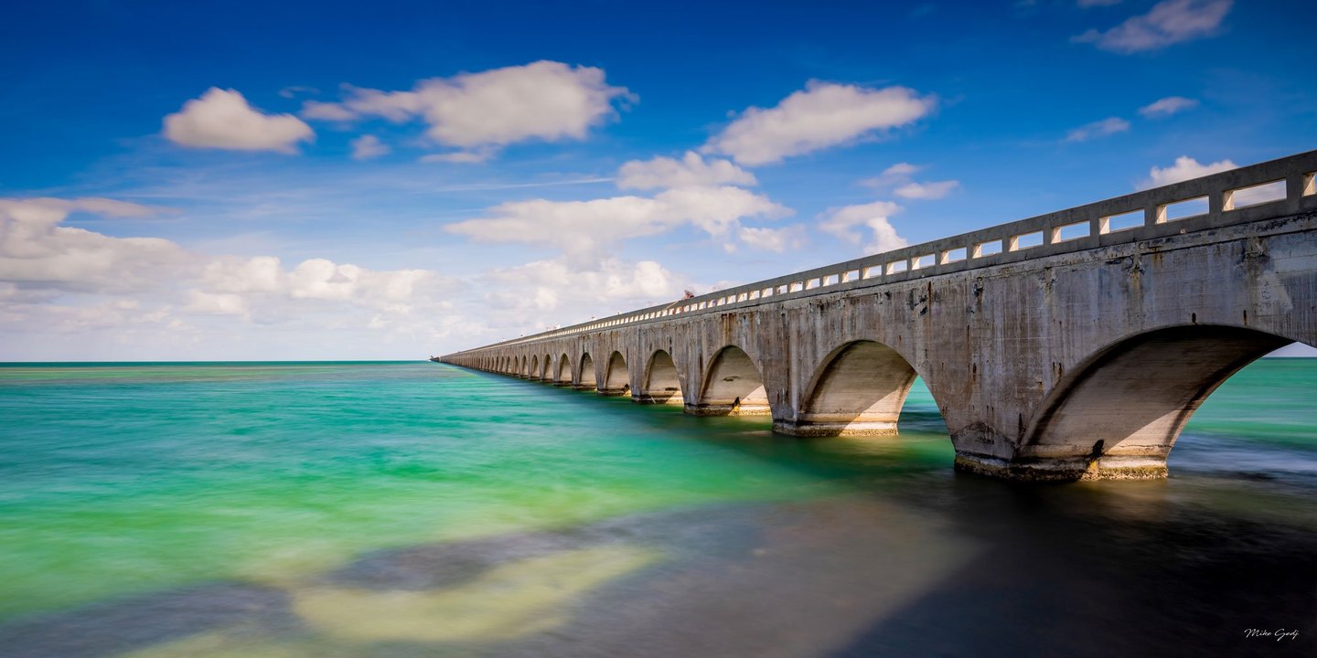 A long concrete bridge with repeating arches stretches toward the horizon over turquoise water beneath a blue sky scattered with white clouds.
