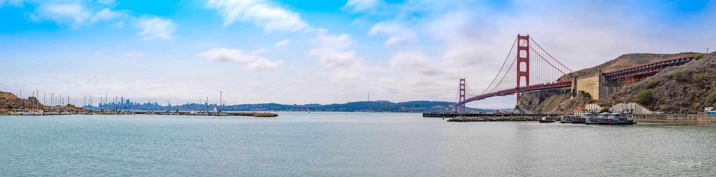 Panoramic view of the Golden Gate Bridge from a sheltered harbor, with sailboat masts on the left, moored vessels on the right, and the distant San Francisco skyline beneath a partly cloudy blue sky.
