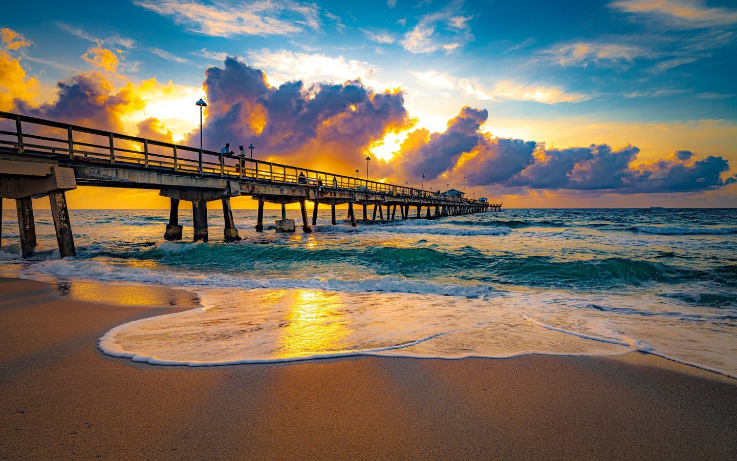 A long pier with scattered people extends over turquoise waves beneath gold-edged clouds, while white foam and sunlight spread across wet sand.