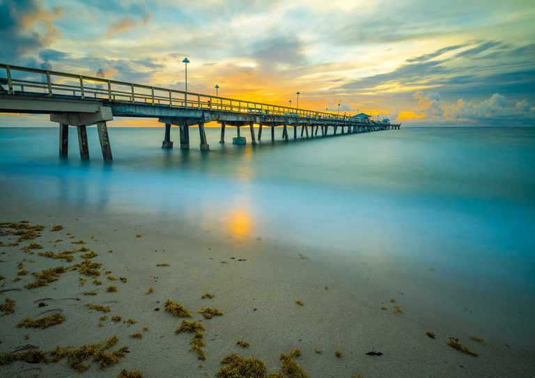 A long concrete pier extends over smooth turquoise water beneath a gold and blue clouded sky, with seaweed scattered across the sandy foreground.