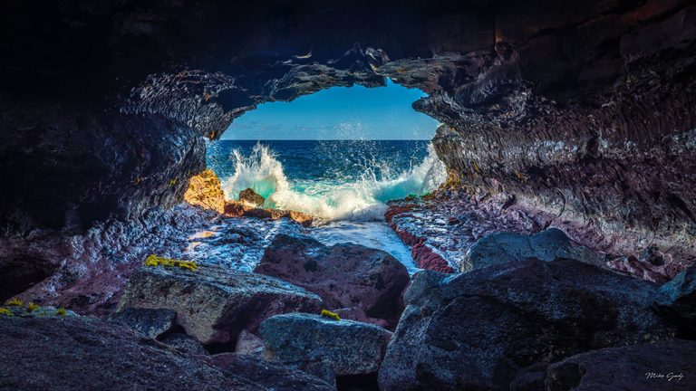 Waves burst against rocks at the opening of a dark sea cave, framing a blue ocean horizon beyond wet foreground boulders.