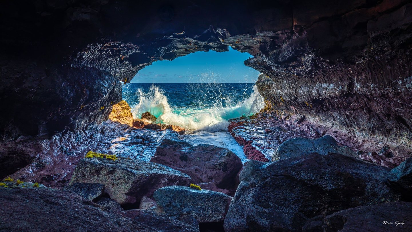 Waves burst against rocks at the opening of a dark sea cave, framing a blue ocean horizon beyond wet foreground boulders.