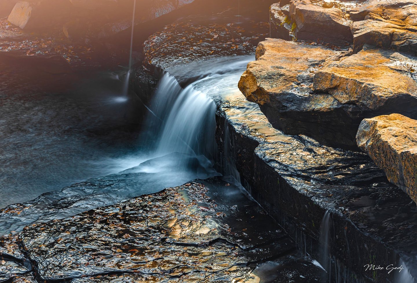 A small waterfall flows over dark, layered rock into a shaded pool beside golden sunlit ledges scattered with fallen leaves.