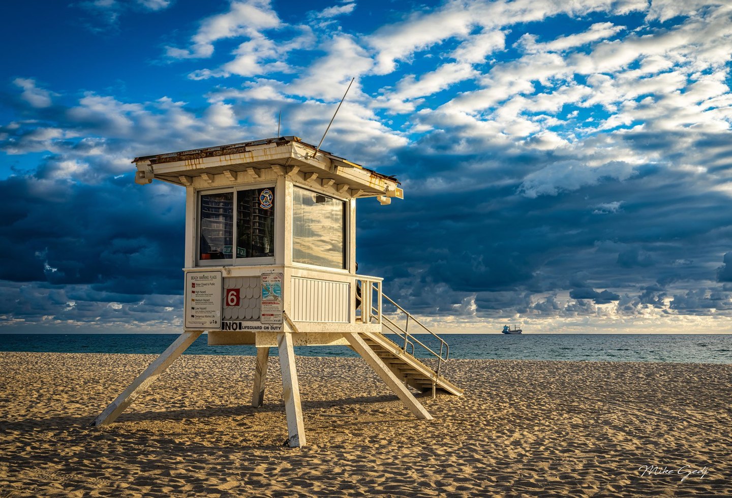 A weathered white lifeguard tower marked 6 stands on an empty sandy beach beneath blue and white clouds, with a ship on the ocean horizon.