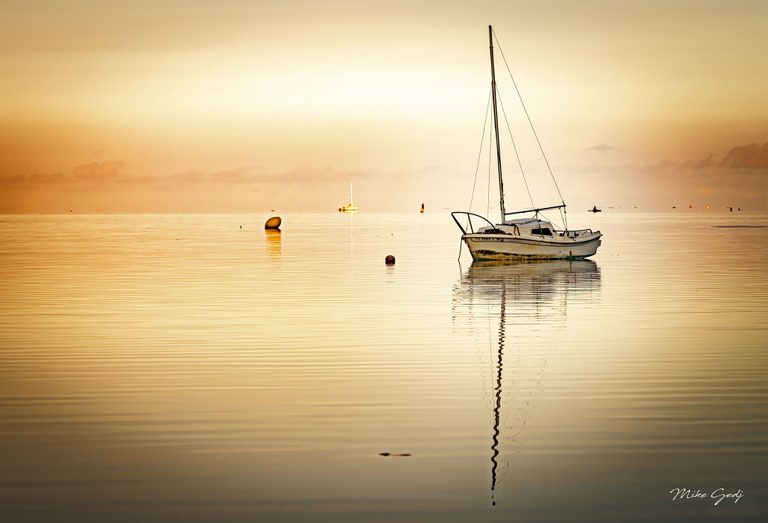 A white sailboat with lowered sails rests on calm golden water, its tall mast reflected among gentle ripples, with buoys and distant boats near the horizon.