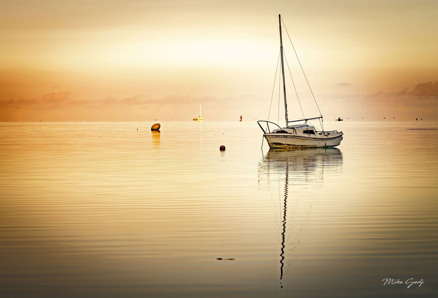 A white sailboat with lowered sails rests on calm golden water, its tall mast reflected among gentle ripples, with buoys and distant boats near the horizon.