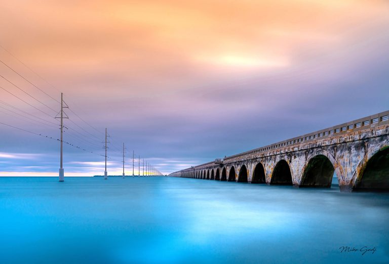 A weathered arched bridge and parallel utility poles recede across turquoise water beneath a peach and lavender sky, with birds perched on the wires.