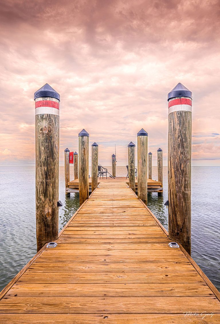 A wooden dock lined with capped timber pilings extends across calm water toward two empty chairs beneath a peach-pink clouded sky.