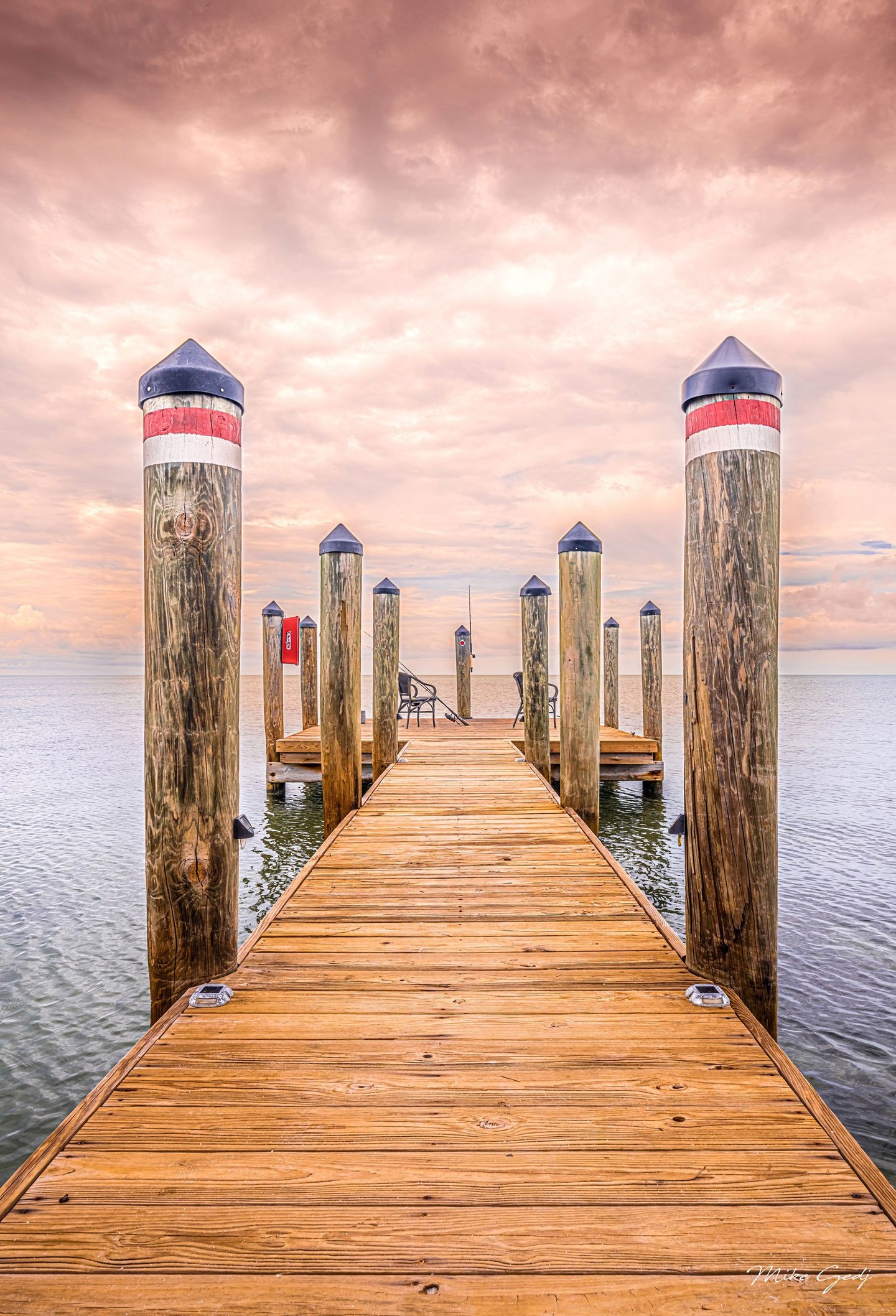 A wooden dock lined with capped timber pilings extends across calm water toward two empty chairs beneath a peach-pink clouded sky.