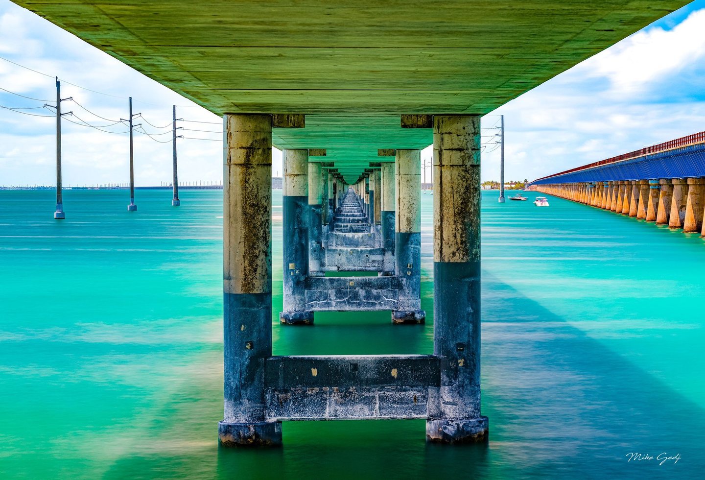 Concrete bridge supports recede symmetrically over turquoise water, with utility poles to the left and a parallel bridge and small boats to the right.