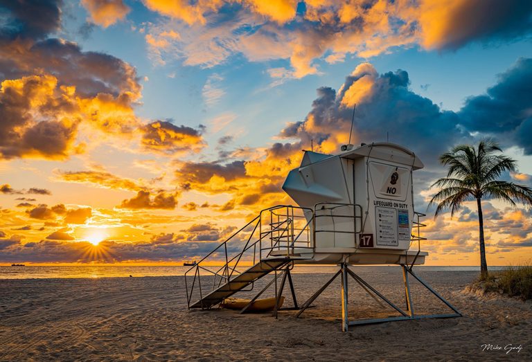 A white lifeguard tower marked 17 stands on a sandy beach beside a palm tree, with the low sun illuminating orange and blue-gray clouds above the ocean.