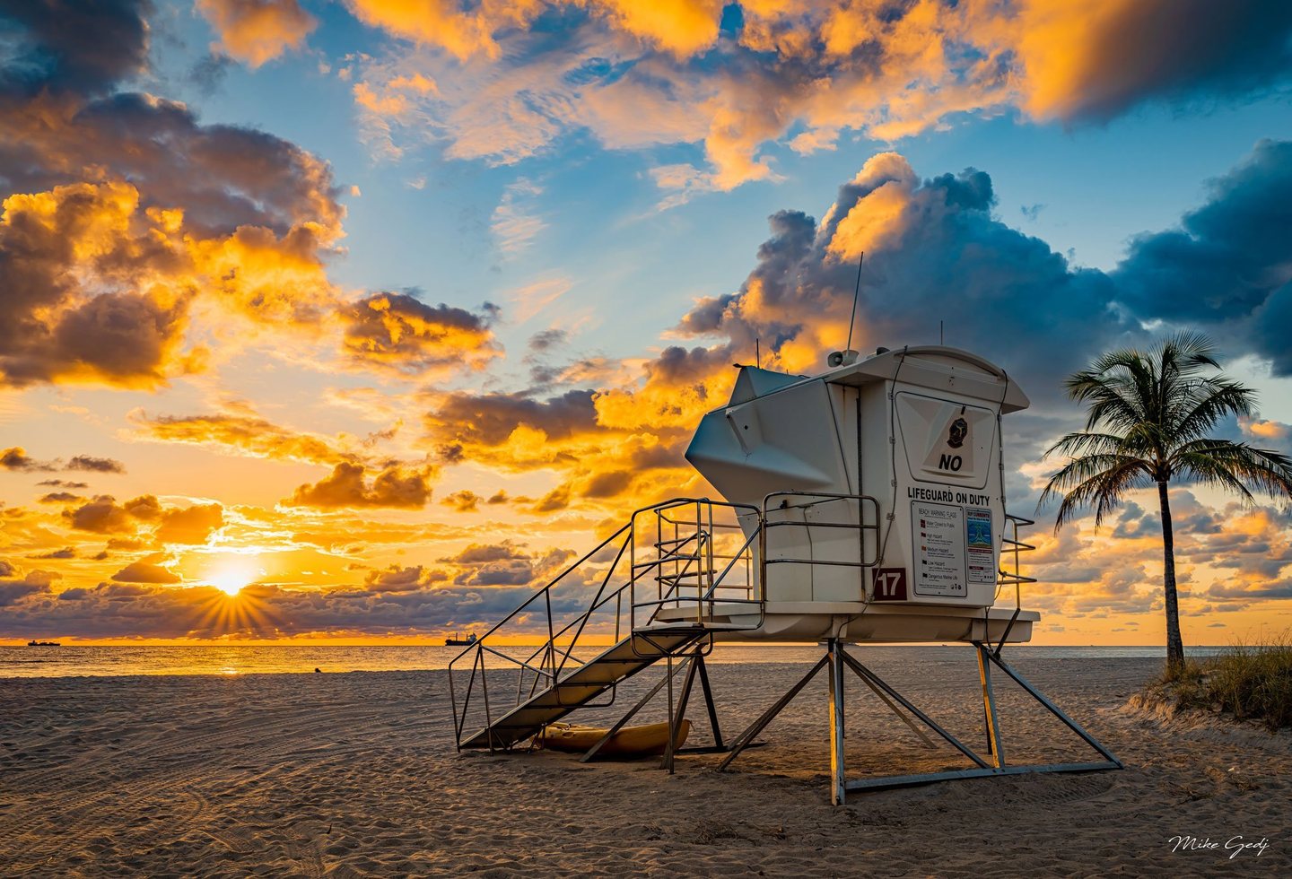 A white lifeguard tower marked 17 stands on a sandy beach beside a palm tree, with the low sun illuminating orange and blue-gray clouds above the ocean.