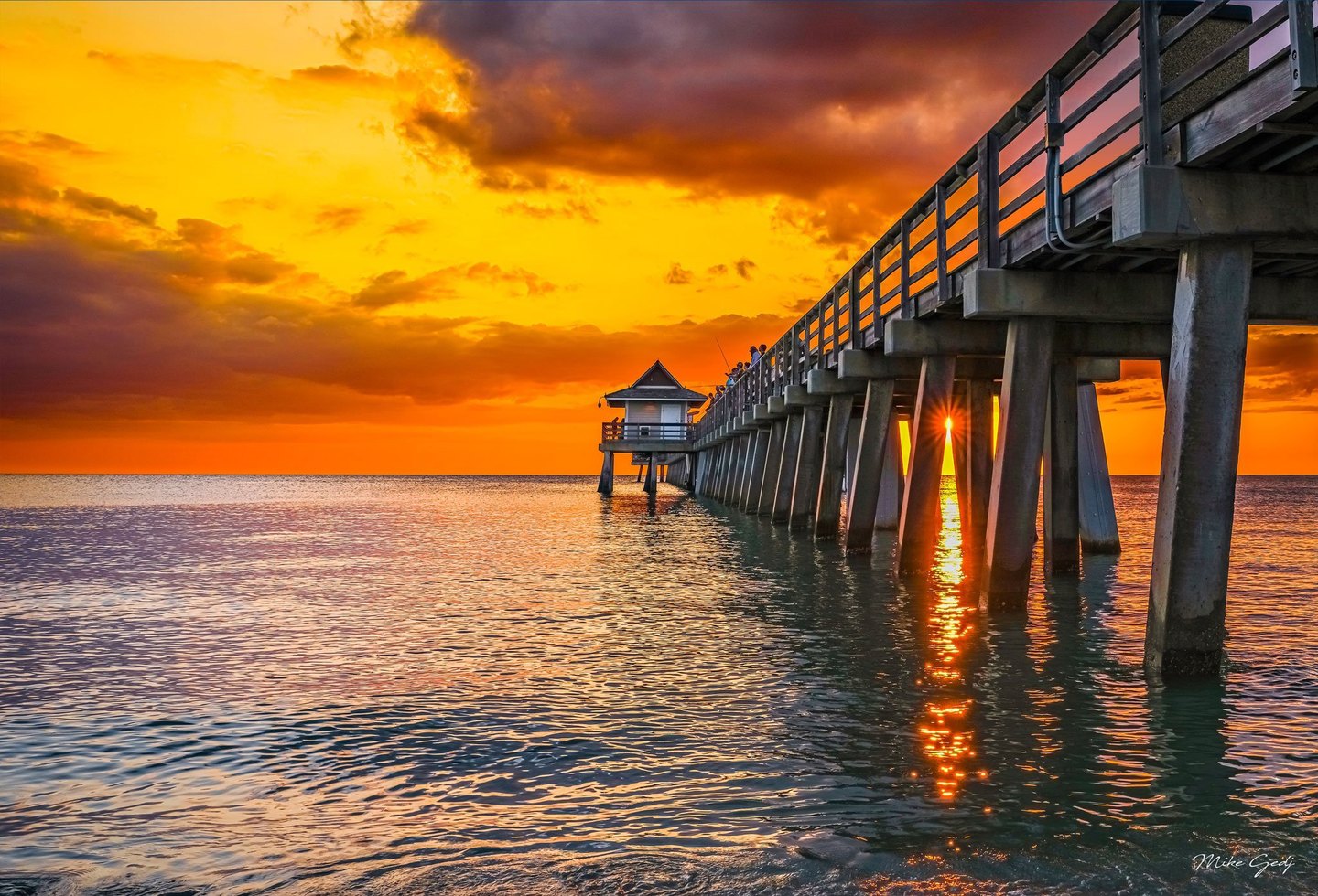 A pier extends over gently rippling seawater beneath an orange sunset sky, with sunlight shining between its concrete pilings and people along its railing.