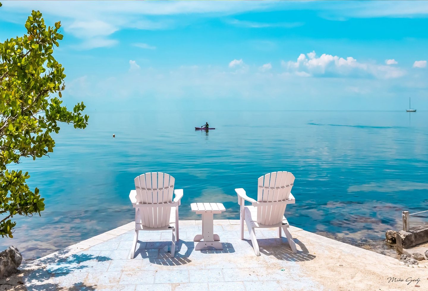 Two white Adirondack chairs and a small table face calm turquoise water from a stone terrace, with leafy branches at left, a lone paddler offshore, and a distant sailboat.