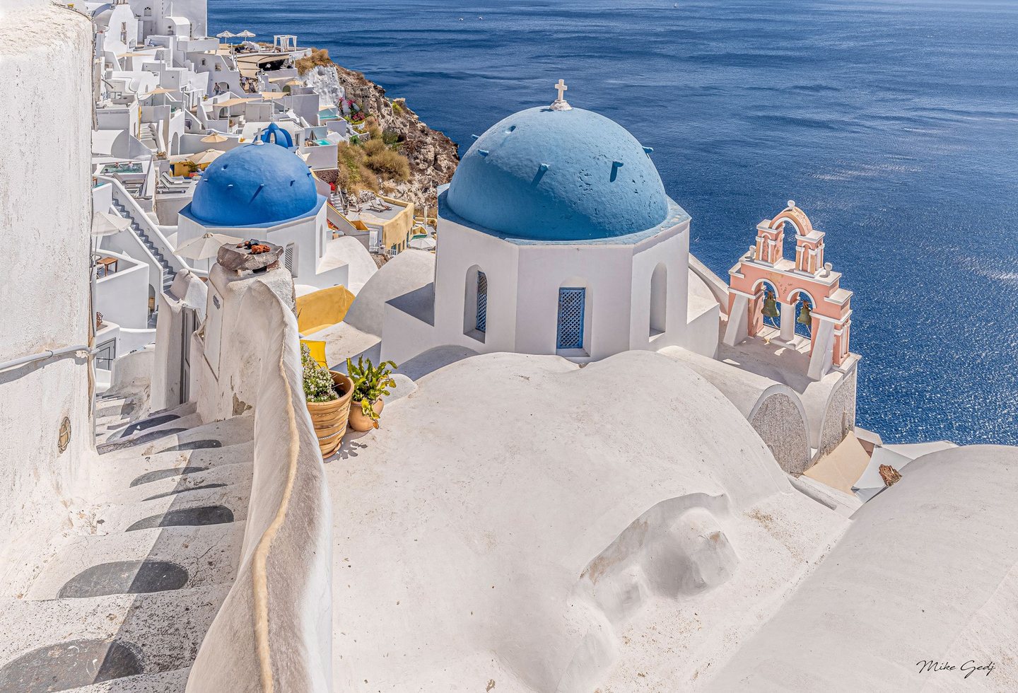 Blue-domed churches and a pink bell tower overlook the deep blue sea in Santorini, with whitewashed rooftops and a narrow stairway in the foreground.