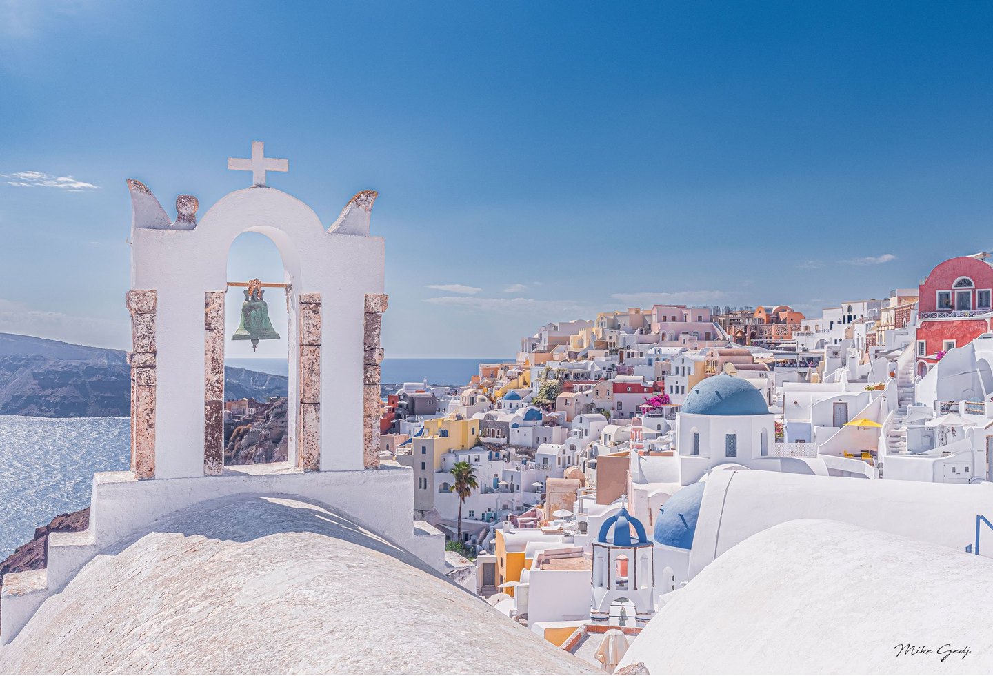 A whitewashed bell tower overlooks blue-domed churches, pastel hillside buildings, and the sunlit sea in Oia, Santorini, beneath a broad blue sky.