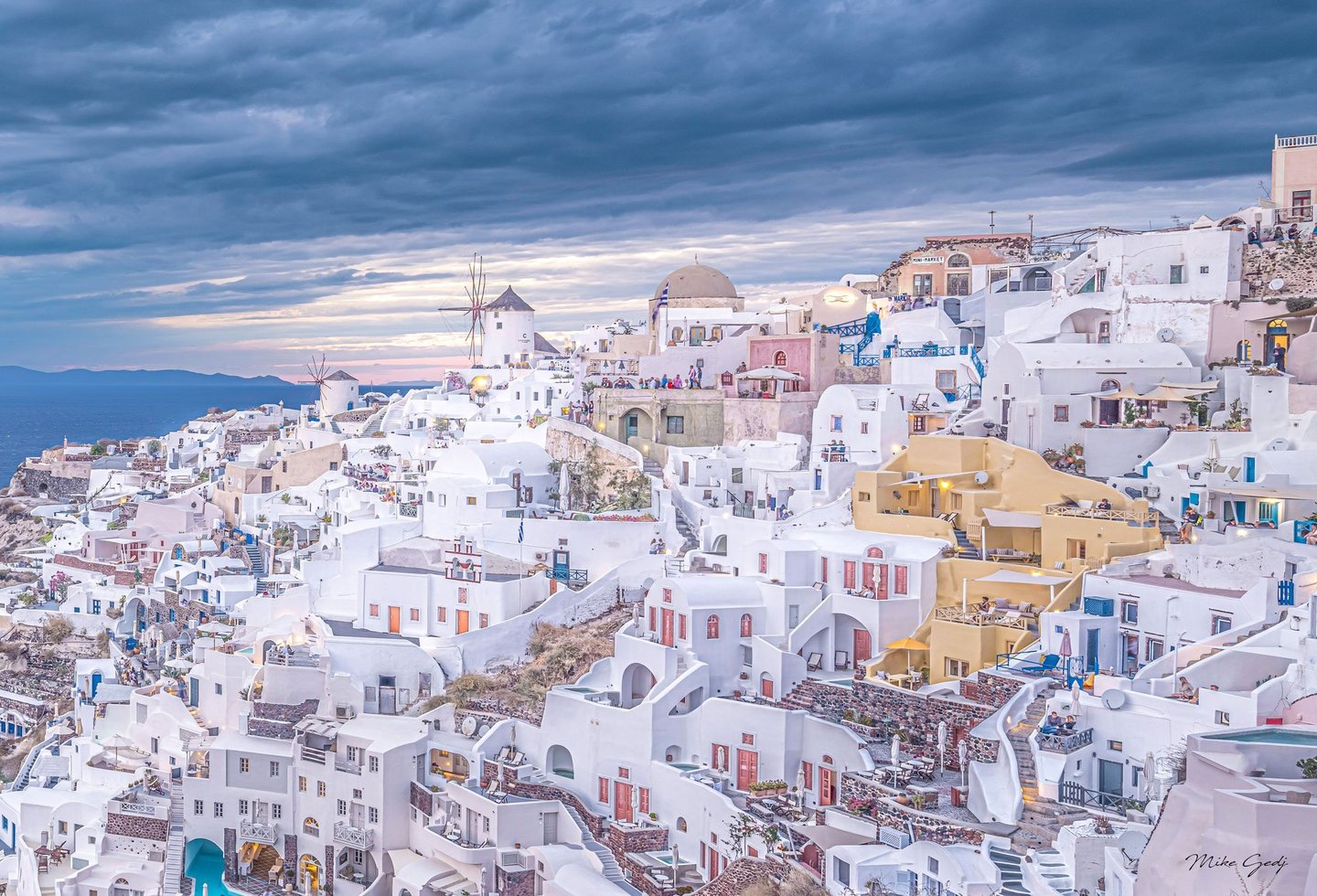 Whitewashed houses, winding stairways, and windmills crowd the hillside of Oia, Santorini, above the blue sea beneath sweeping evening clouds.