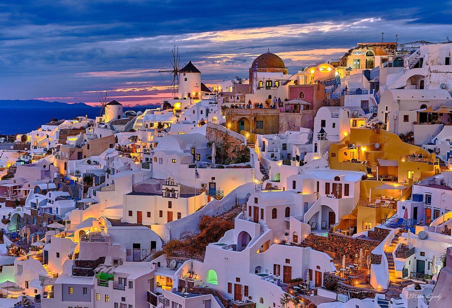 Whitewashed buildings, windmills, and winding stairways in Oia, Santorini, glow with warm lights beneath a blue and pink twilight sky above the sea.