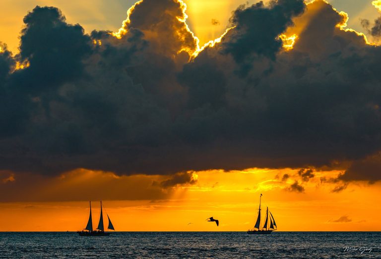 Two silhouetted sailboats and flying seabirds sit beneath towering dark clouds edged with golden sunlight over an orange sea horizon.