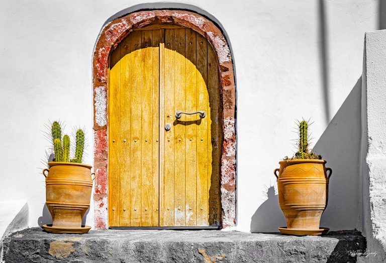 A yellow wooden door beneath a reddish stone arch stands in a white plaster wall, flanked by cacti in tall terracotta pots under strong sunlight.