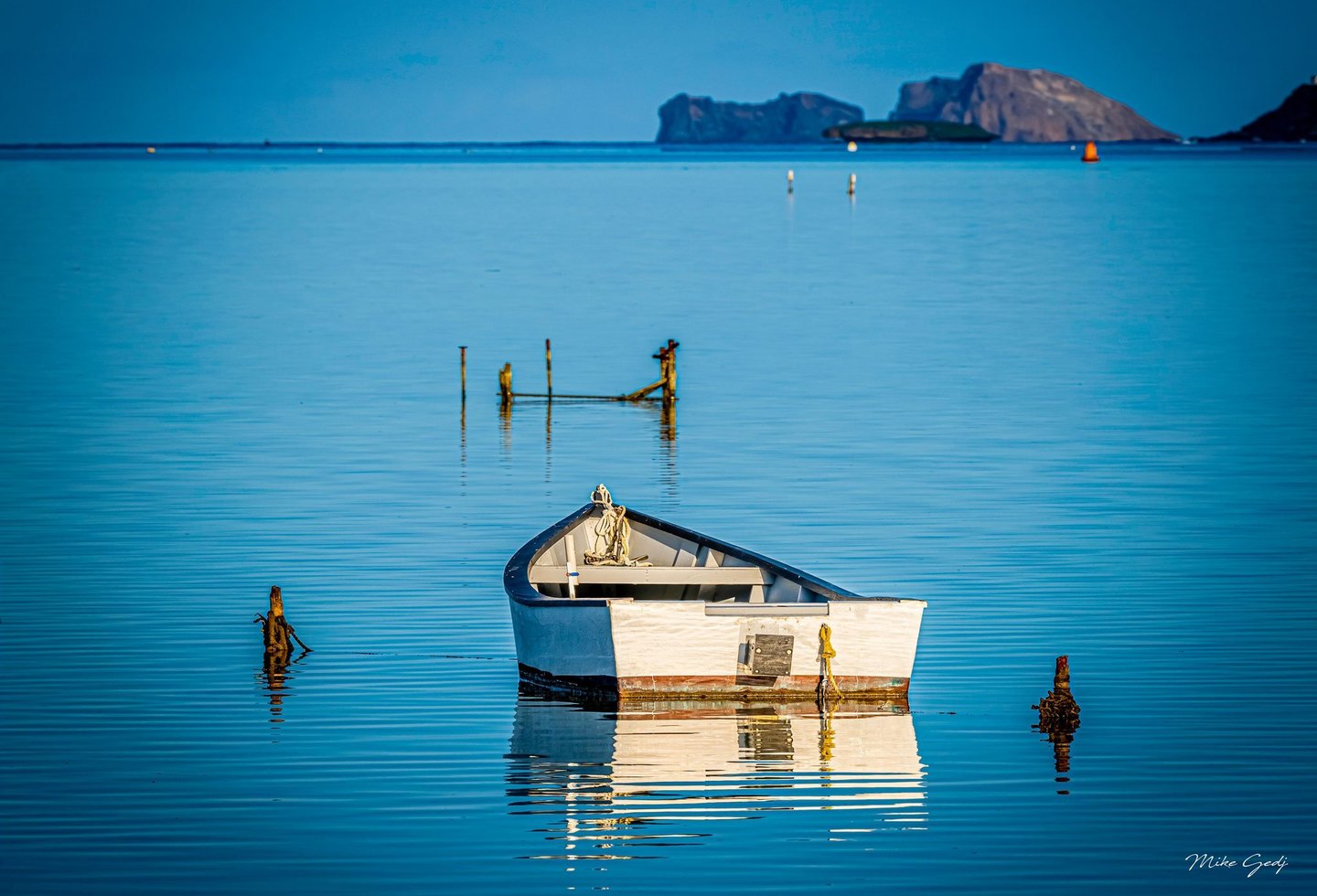 A small white boat floats among weathered posts on calm blue water, its reflection rippling beneath it, with buoys and rocky islands in the distance.