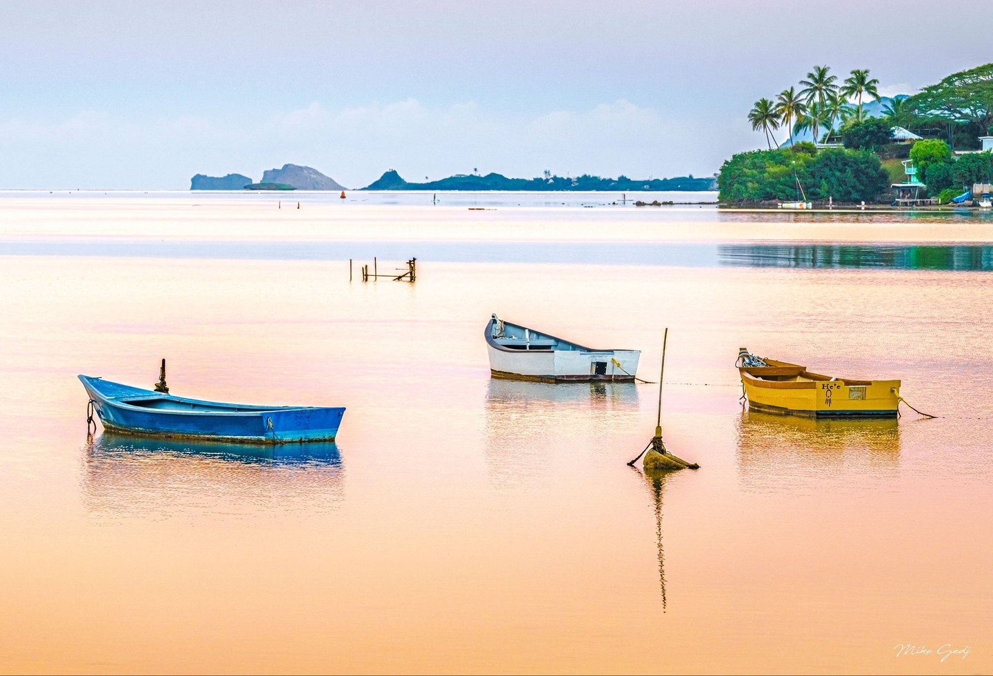 Blue, white, and yellow boats sit moored in calm peach-colored water, with distant rocky islands and a palm-lined shoreline beneath a pale blue sky.