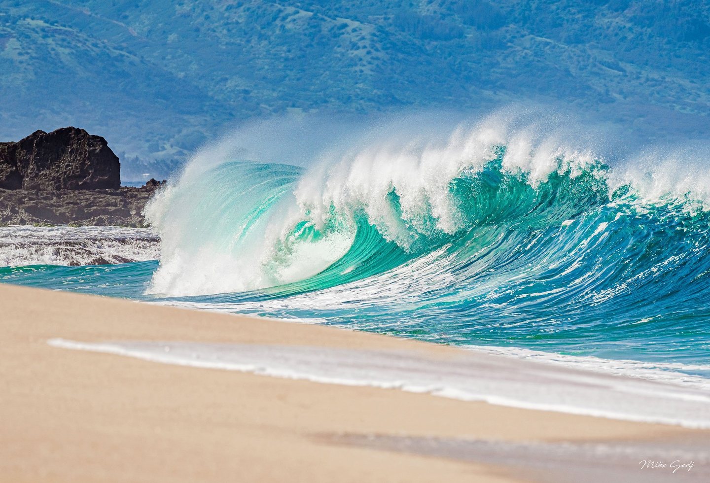 A turquoise wave breaks beside a sandy beach, throwing white spray into the air with dark coastal rocks and hazy green hills behind it.