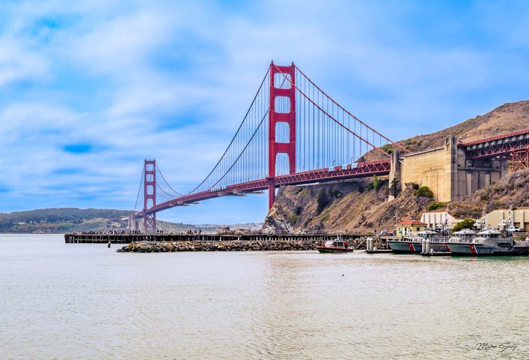 The Golden Gate Bridge spans the water beneath a cloud-streaked blue sky, with dry hills, a waterfront pier, and moored Coast Guard boats in the foreground.