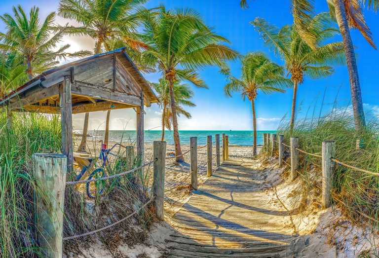 A sand-covered wooden boardwalk with rope railings leads past a weathered shelter and palm trees to a turquoise sea beneath a bright blue sky.