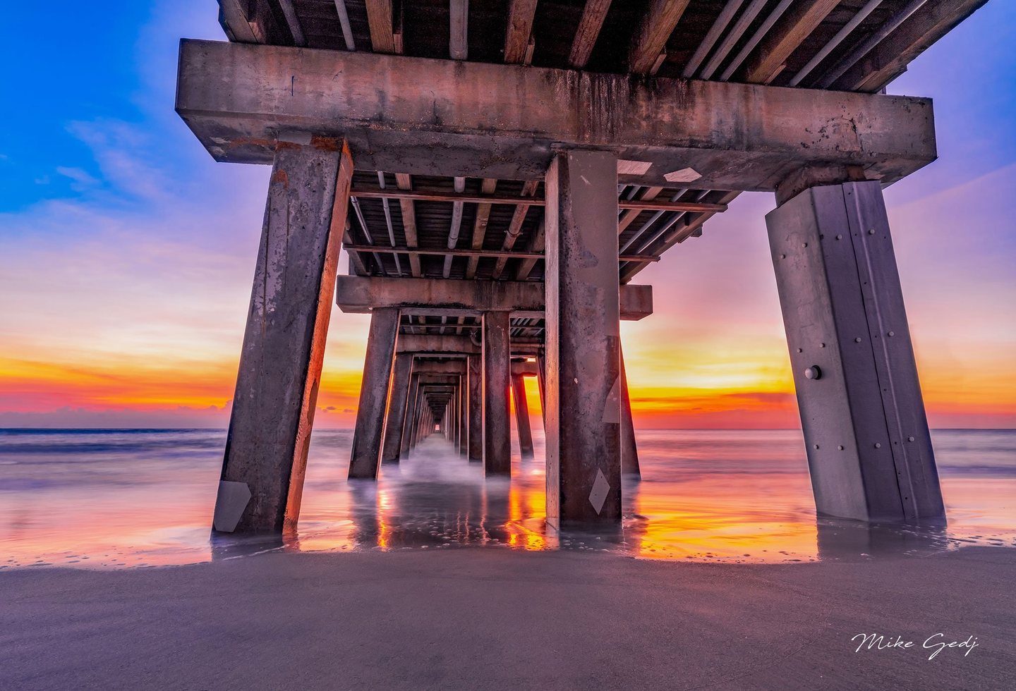 Concrete pier supports recede toward the ocean beneath a blue, pink, and orange sky, with softened surf and bright reflections across wet sand.