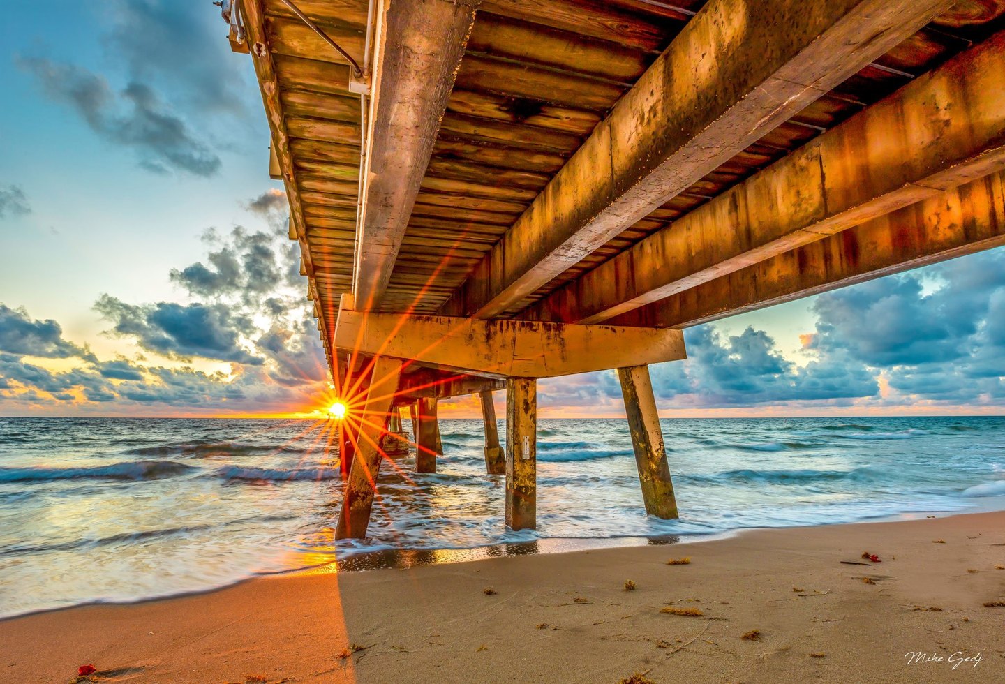 Low sunlight forms a bright starburst beneath a weathered pier as waves wash onto a sandy beach under a blue, cloud-filled sky.