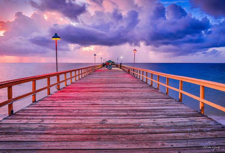A wooden pier lined with glowing lamps extends toward distant people and a small pavilion over the ocean beneath pink, purple, and blue clouds.