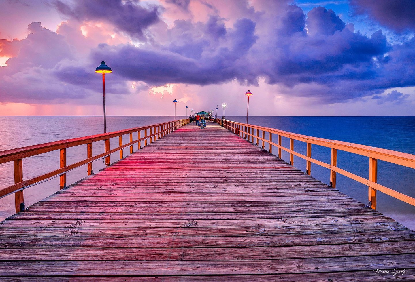 A wooden pier lined with glowing lamps extends toward distant people and a small pavilion over the ocean beneath pink, purple, and blue clouds.