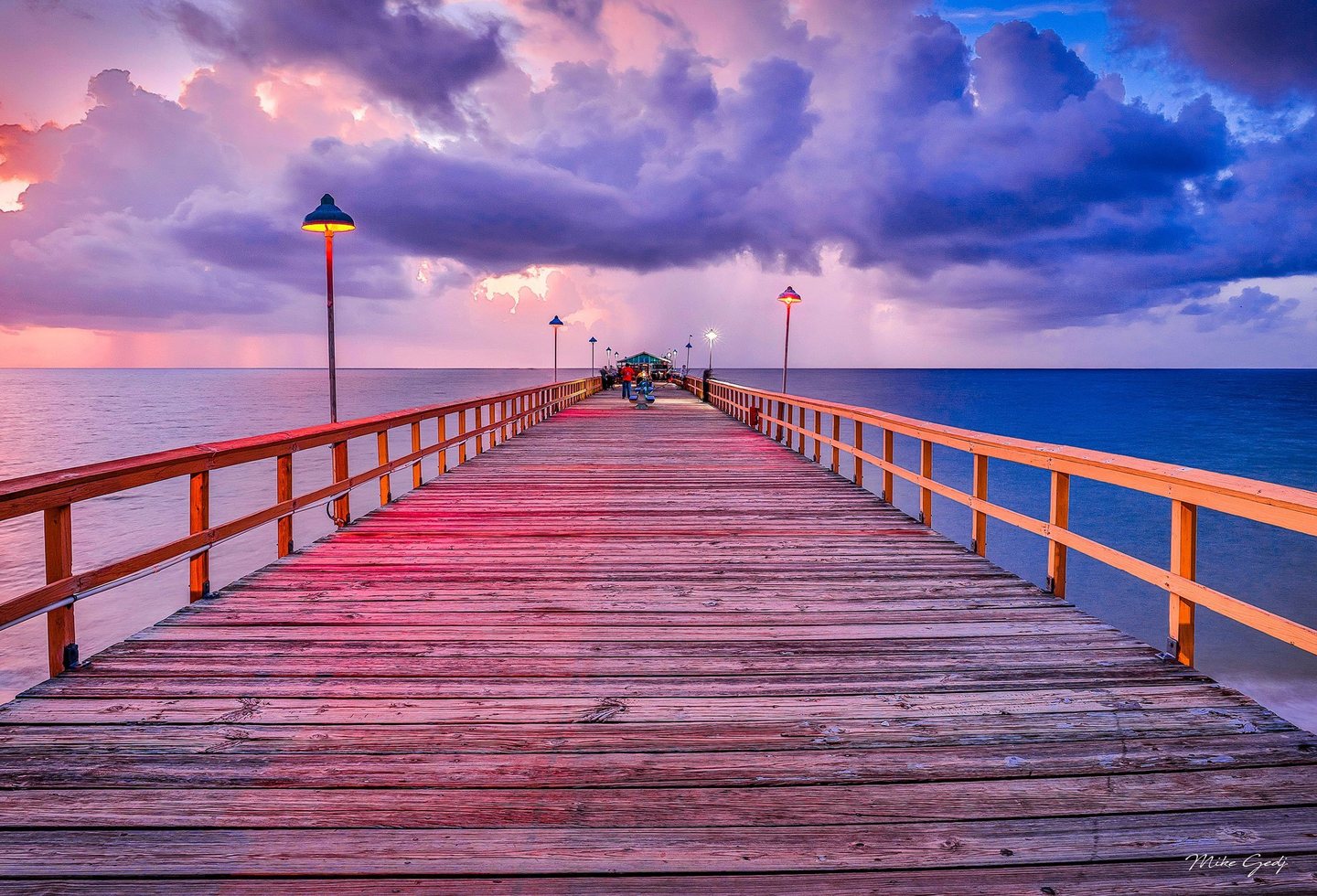 A wooden pier lined with glowing lamps extends toward distant people and a small pavilion over the ocean beneath pink, purple, and blue clouds.