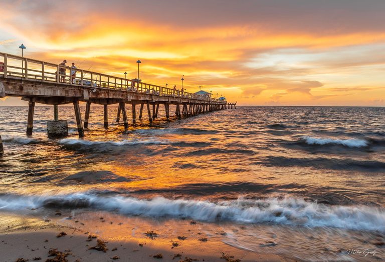 People fish from a long pier extending over the ocean beneath an orange and gold sky as foamy waves wash onto seaweed-strewn sand.