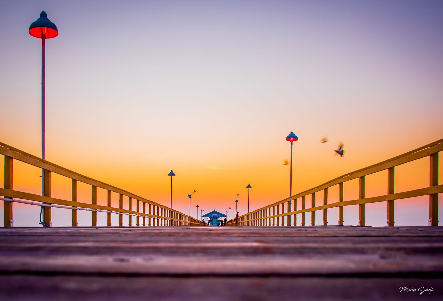 A low view along a wooden pier shows converging yellow railings, glowing red lamps, distant people, and birds flying across an orange and lavender sky.