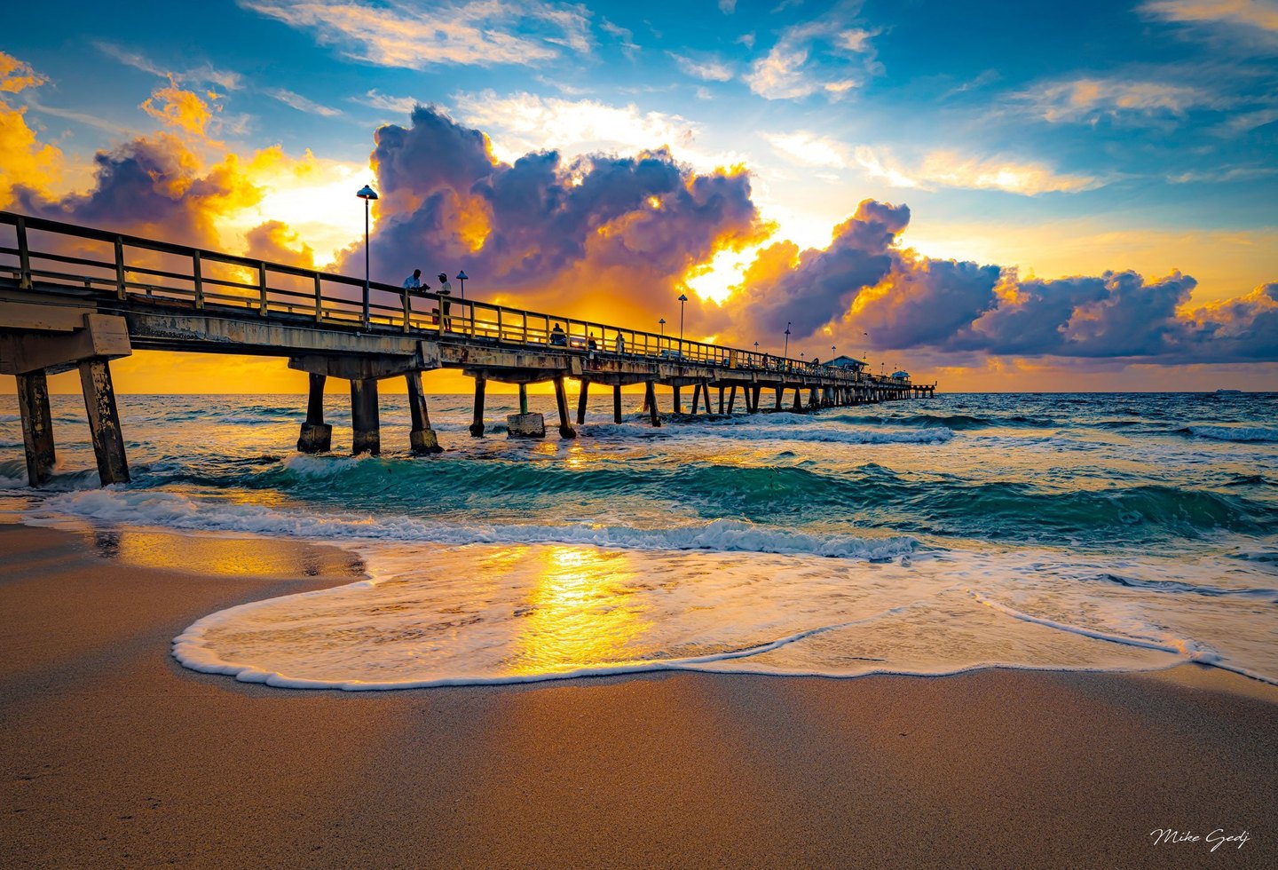 A long pier with scattered people extends over turquoise waves beneath golden backlit clouds, while sea foam and sunlight spread across wet sand.