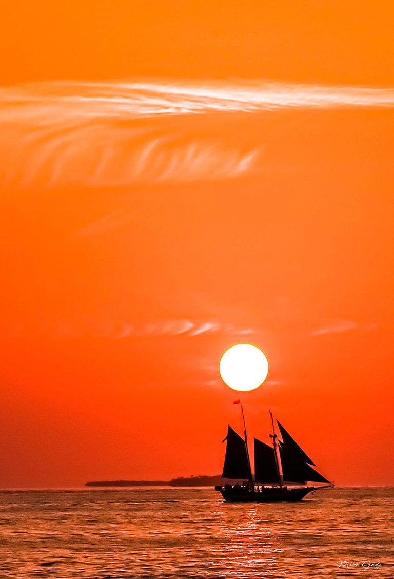 A silhouetted two-masted sailboat crosses rippling water beneath a bright sun in a vivid orange sky streaked with thin white clouds.