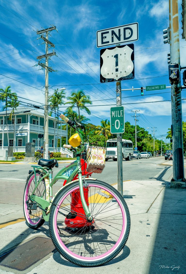 A mint-green bicycle with pink wheel rims stands beside a red fire hydrant beneath the US Route 1 end and mile zero signs in sunny Key West.