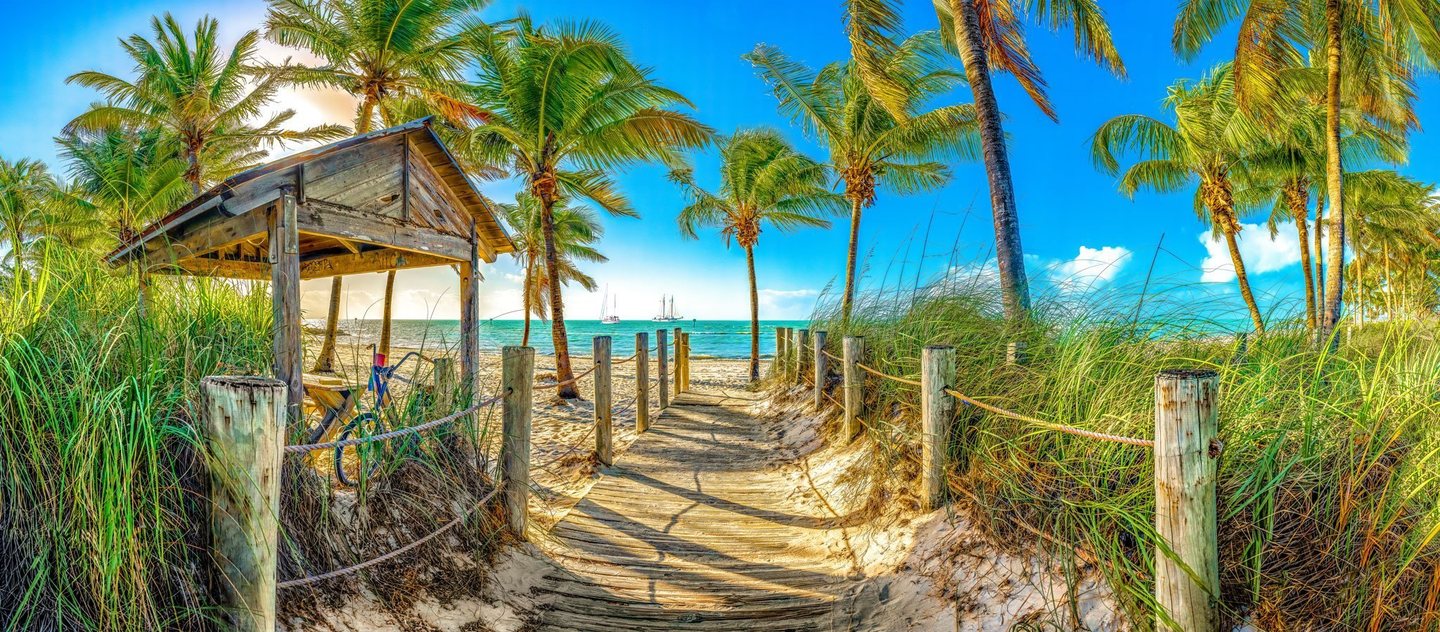 A rope-lined wooden boardwalk leads through dune grasses and palm trees past a weathered shelter to a turquoise sea with distant sailboats.