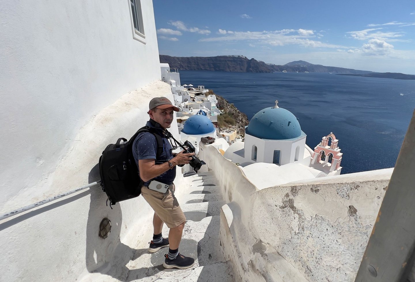 A man holding a camera pauses on a narrow whitewashed stairway above blue-domed churches and the deep blue sea in Santorini.