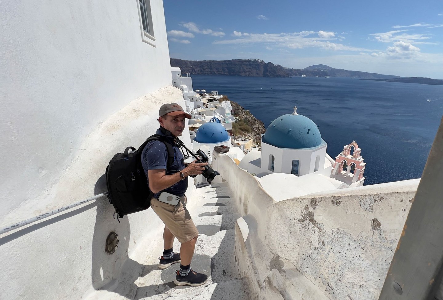 A man holding a camera pauses on a narrow whitewashed stairway above blue-domed churches and the deep blue sea in Santorini.