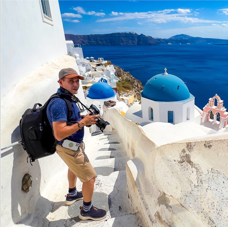 A man holding a camera stands on a narrow whitewashed stairway above blue-domed churches and the deep blue sea in Santorini.